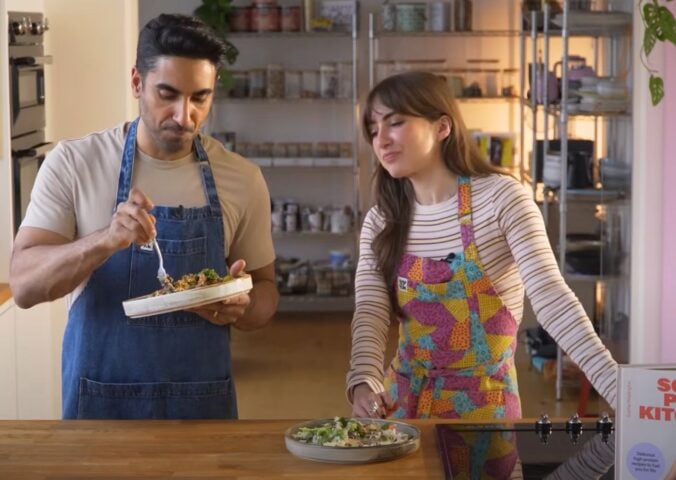 Dr. Rupy Aujla and Sophie Waplington stand by a counter cooking high-protein vegan work lunches